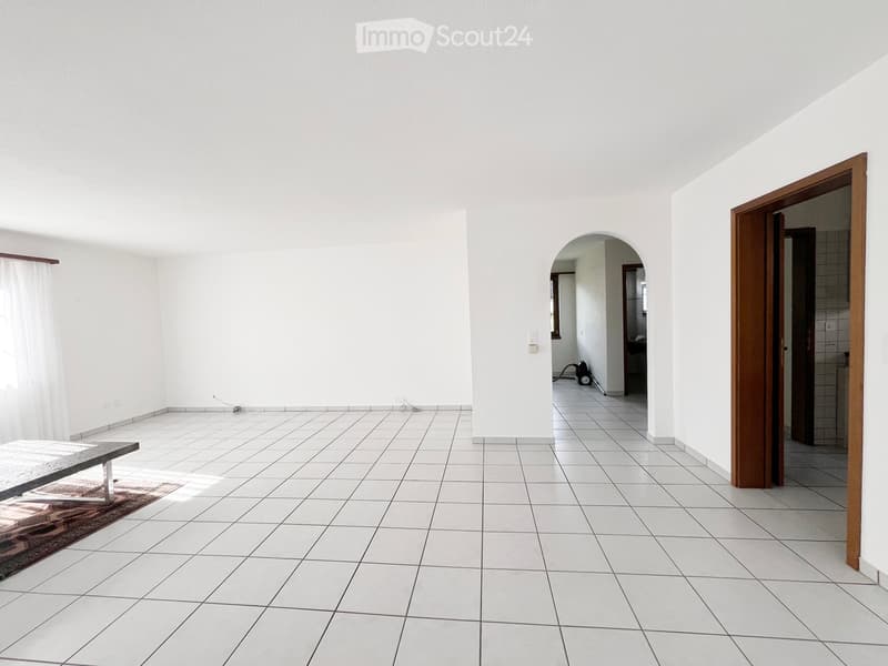 Bright, unfurnished room with white tiled flooring, white walls, and a wooden bench with a rug placed on the floor. An arched doorway leads to another room.