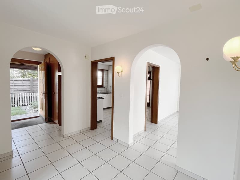 Bright hallway with white tiled flooring, arched doorways leading to other rooms, and a view of an outdoor area through an open door.