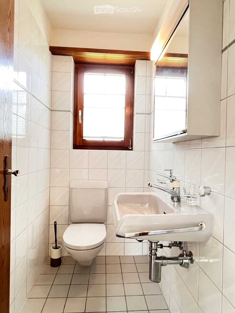 A small bathroom with white tiled walls and floor, featuring a toilet, sink with a faucet, a mirror above the sink, and a small window with wooden frame.