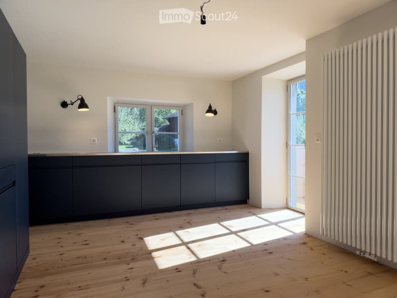 Empty kitchen with dark grey cabinets, light wooden floor, and large windows allowing natural light to enter. There is a radiator on the right wall and wall-mounted lights on either side of the window.