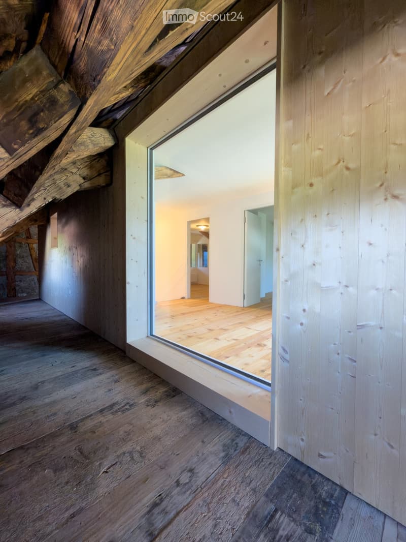 A modern hallway with wooden floors, exposed wooden beams, and large glass windows leading to another room.