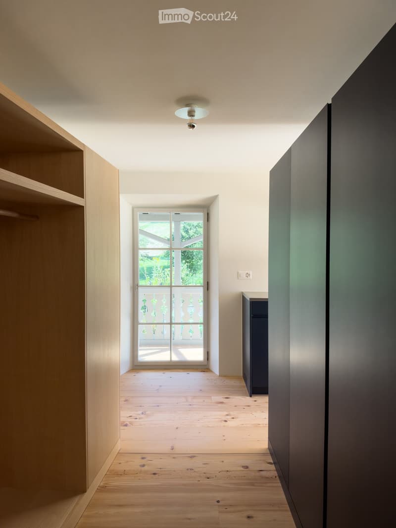 Empty hallway with wooden flooring, white walls, and a large window at the end leading to an outdoor area.