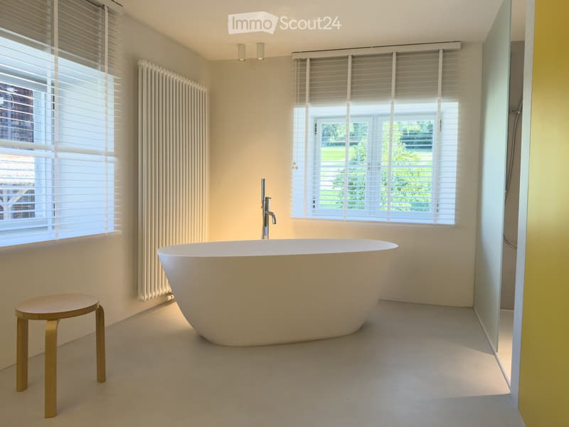Modern bathroom with a freestanding bathtub, a small wooden stool, a modern faucet, and a radiator near the window.