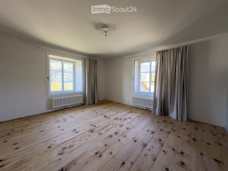 Empty room with wooden flooring, large windows with curtains, radiators, and a ceiling light fixture.