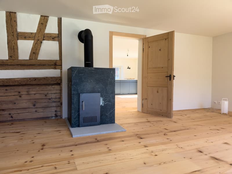 Empty room with wooden flooring, exposed wooden beams, and a modern black wood-burning stove with a chimney. A wooden door leads to another room.