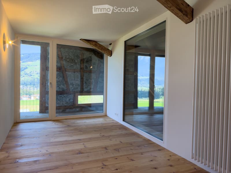 Empty room with wooden flooring, large glass doors leading to a balcony, white walls, and a view of the countryside.