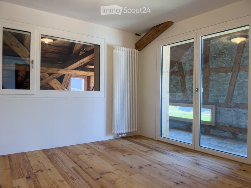 Empty room with wooden flooring, white walls, exposed wooden beams, large windows, and a radiator. The room has glass sliding doors leading to an outdoor area.