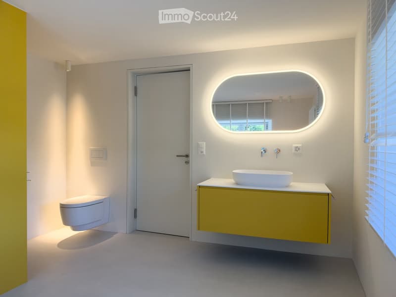 Modern bathroom with white walls, a floating yellow cabinet, a white sink, a toilet, and an illuminated oval mirror above the sink.