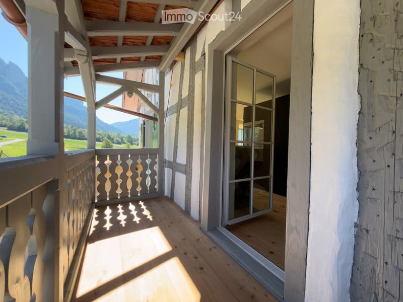 Wooden balcony with ornate railing, overlooking a scenic valley and mountains, with large glass doors leading inside.