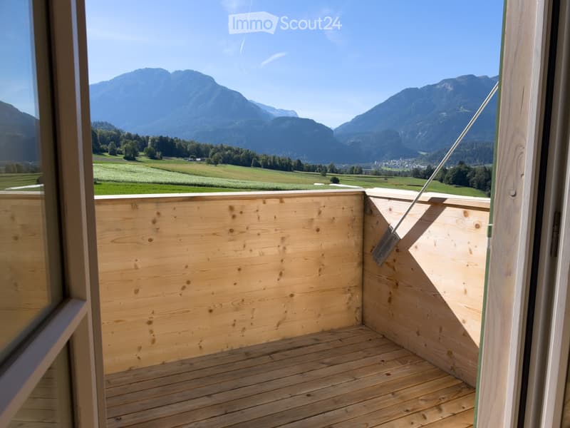 Wooden balcony with open sides, overlooking a scenic mountain landscape and green fields.
