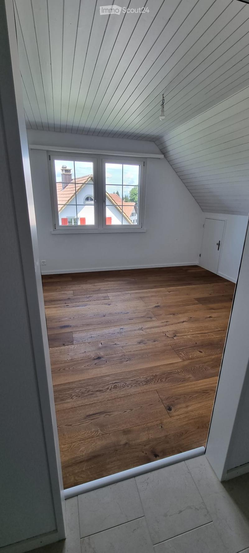 Empty attic room with wooden flooring, white walls, and a large window offering a view of the outside. The ceiling has a sloped design with exposed wooden beams.