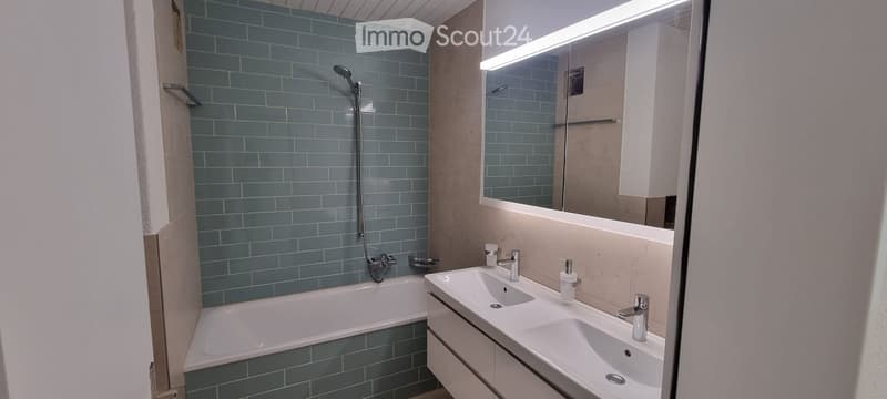 Modern bathroom with blue tiled walls, a bathtub, double sink vanity, and a large mirror with built-in lighting.