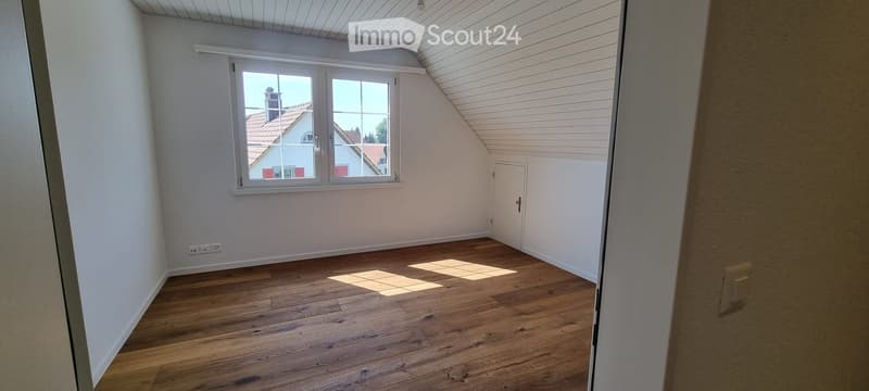 Empty attic room with wooden flooring, white walls, and a large window offering a view of a neighboring house with a red roof.