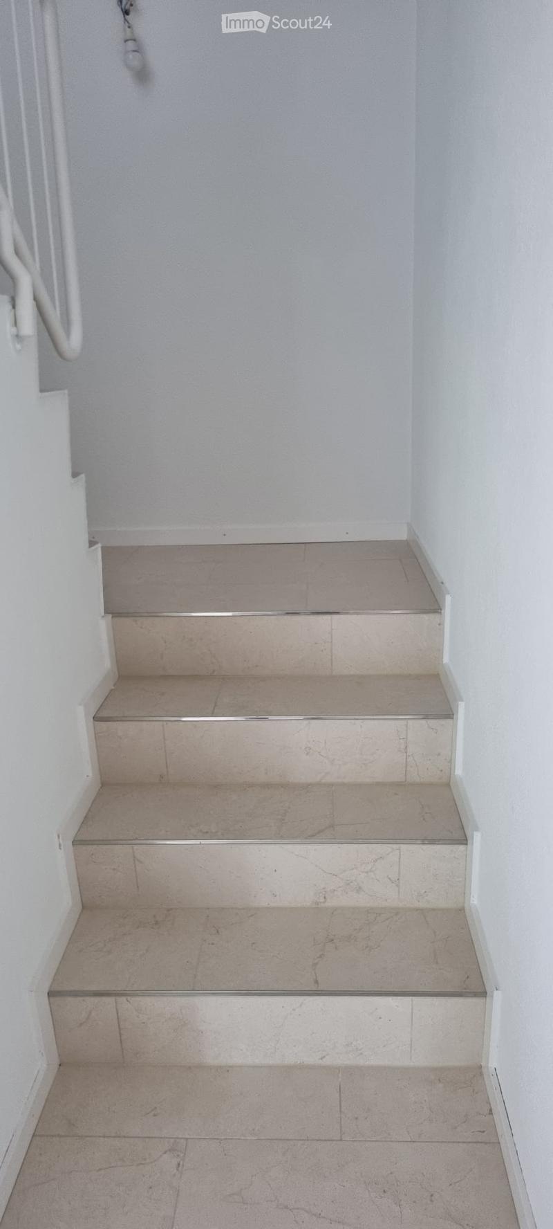 A short staircase with beige tiled steps and white walls, featuring a handrail on one side.
