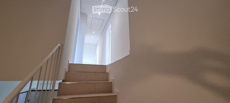 Staircase with beige steps and a wooden handrail, leading to a white ceiling with recessed lighting. The walls are painted in a warm, earthy tone, and the space is well-lit with natural light coming from the left side.