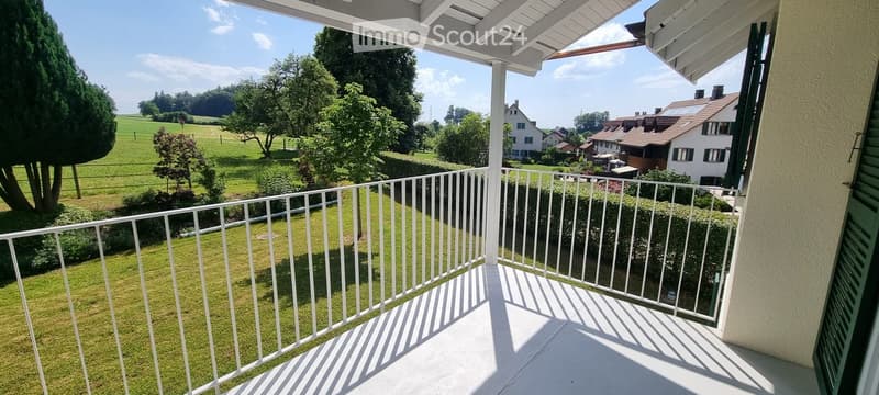 Balcony with white railing, overlooking a green lawn and countryside with trees and houses in the distance.