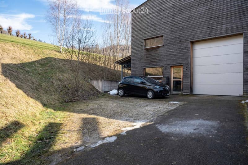 Modern wooden building with a garage, situated on a raised ground floor with a sloped driveway leading up to it. A black car is parked in front of the garage.