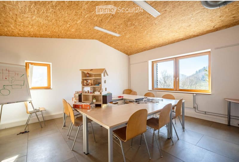 Bright room with wooden ceiling, large windows, a long table surrounded by chairs, a small shelf with various items, and a whiteboard on the wall.