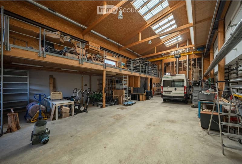 Large, open workshop or garage space with wooden beams, concrete floor, and scattered tools and equipment. Features a loft area with a metal railing, a van parked inside, and various storage items and machinery.