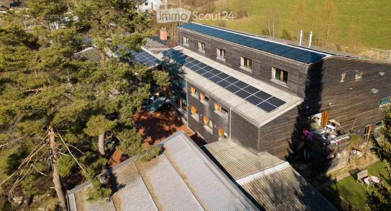 A modern, multi-story house with dark wooden exterior and solar panels on the roof, surrounded by greenery and trees.