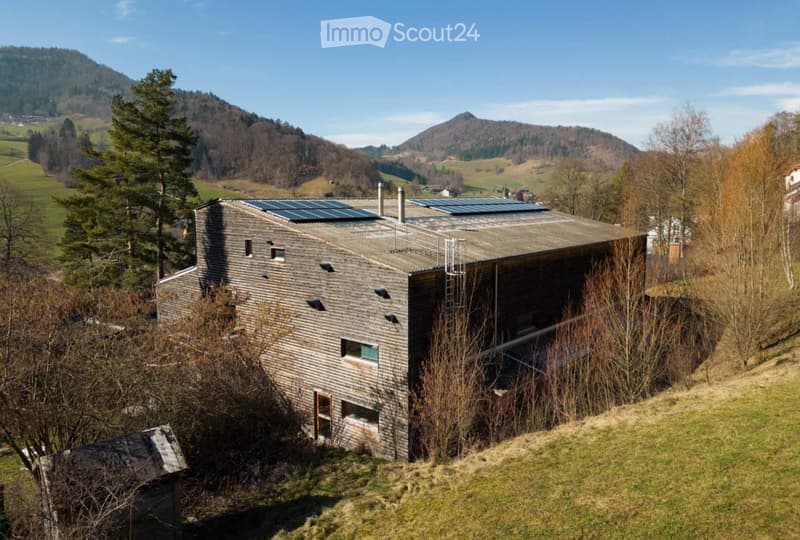 A modern wooden house with a flat roof and solar panels, situated in a rural landscape with trees and hills in the background.