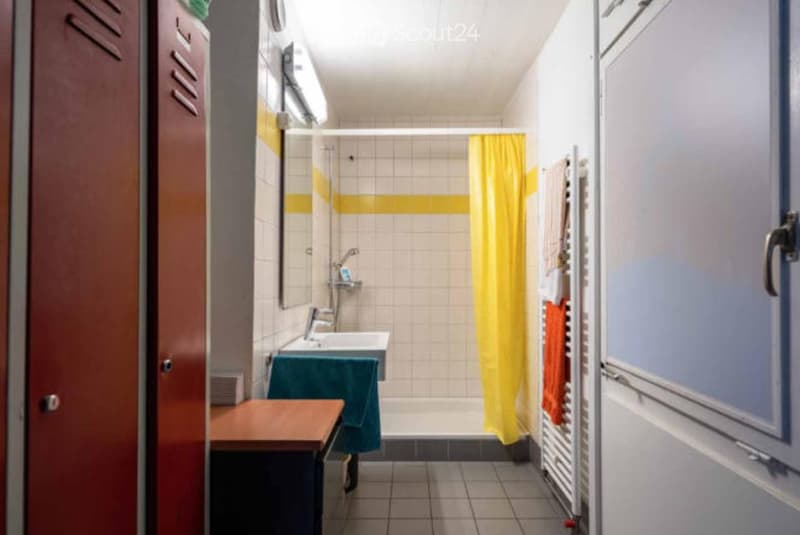 A compact bathroom with a sink, mirror, and shower area. The shower has a yellow curtain, and there is a towel rack with towels on the right side. The walls are tiled with a yellow accent stripe.