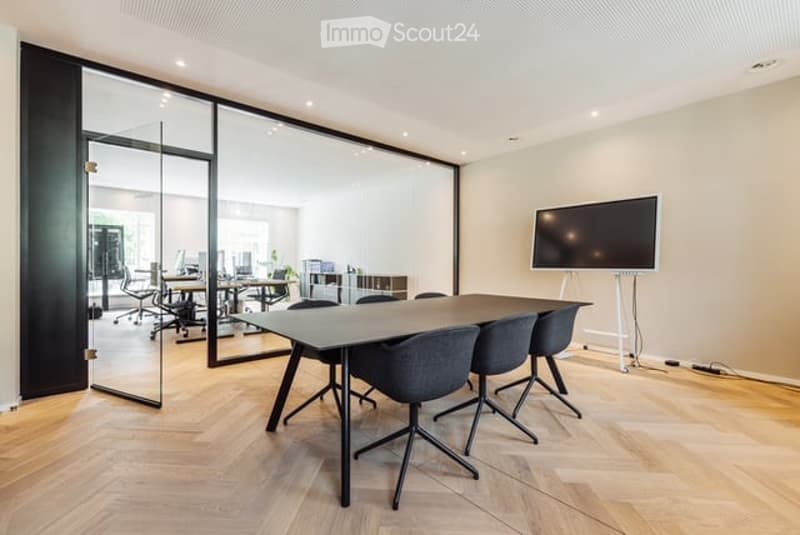 Modern conference room with a large wooden table, several chairs, and a large screen on the wall. The room features large glass walls and a sleek, contemporary design.