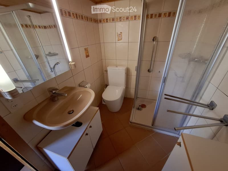Bathroom with a sink, toilet, and a walk-in shower with grab bars. The walls are tiled with a decorative border, and the floor is tiled in a light brown color.