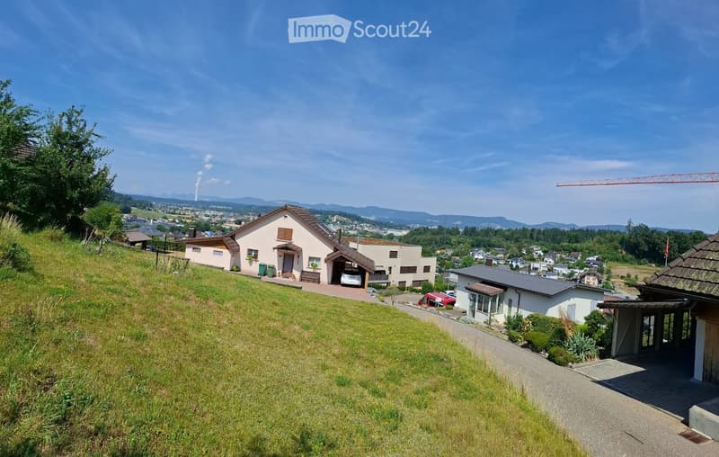 A two-story house with a garage, situated on a hillside with a view of a town and surrounding mountains. The house has a light-colored exterior with a brown roof and is surrounded by greenery.