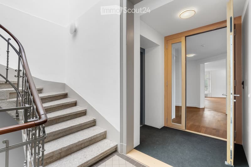 Modern hallway with a staircase featuring a wooden handrail and metal balustrade, leading to an open wooden door that reveals a room with wooden flooring and large windows.