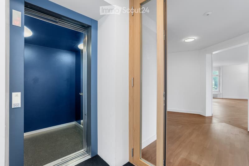 Modern hallway with white walls, wooden flooring, and a glass elevator door on the left. The hallway features recessed lighting and a visible room with wooden flooring and large windows at the end.