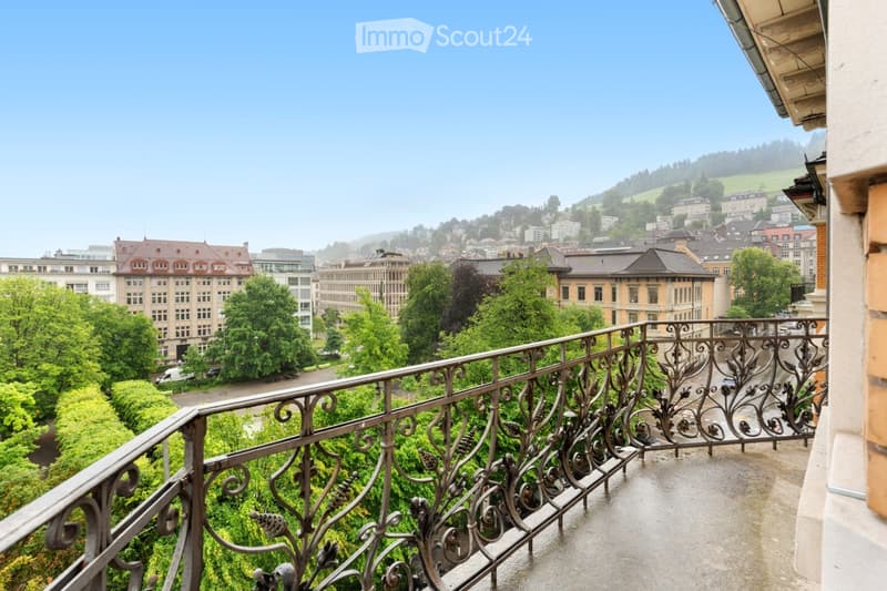 Balcony with ornate metal railing overlooking a cityscape with buildings, trees, and distant hills.