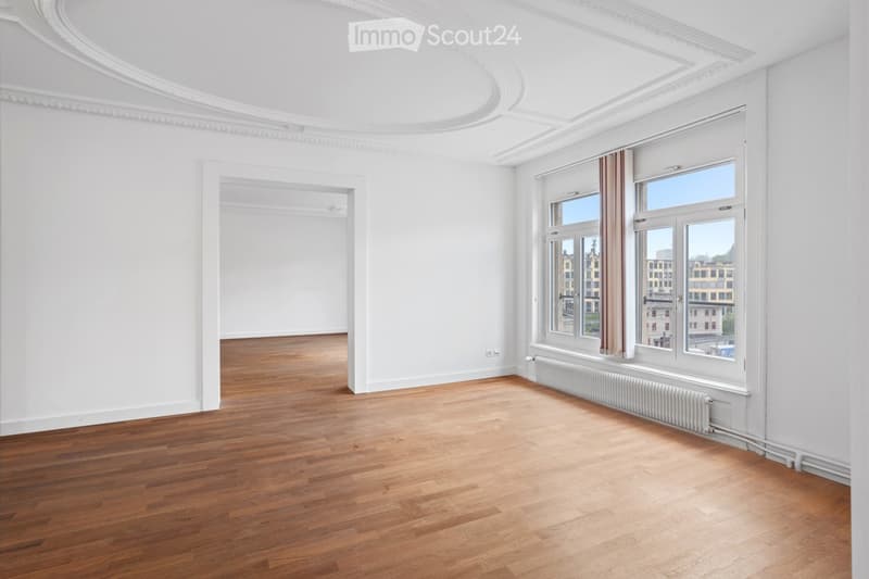 Empty room with wooden flooring, white walls, large windows with curtains, and a radiator under the windows.