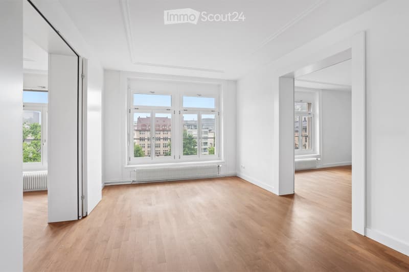 Completely empty room with white walls, wooden floor, large windows, and radiators. No furniture or equipment.