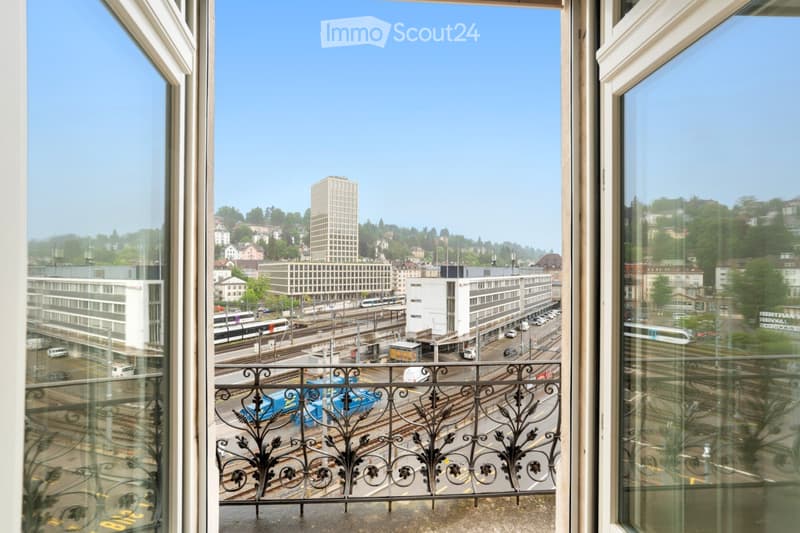 Balcony overlooking a cityscape with buildings, train tracks, and traffic, featuring a decorative metal railing.