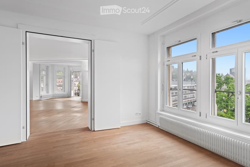 Bright, unfurnished room with wooden flooring, white walls, large windows, and a radiator. The room is empty, with no furniture or personal items visible.