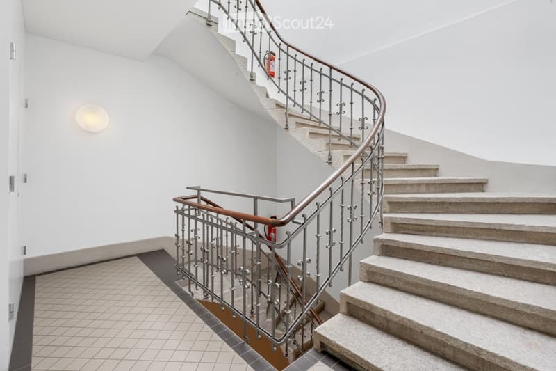 Spiral staircase with metal railing and beige steps, white walls, and a wall-mounted light fixture.