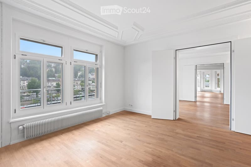 Bright, unfurnished room with white walls, wooden flooring, large windows offering a view of the city, and a radiator below the windows.