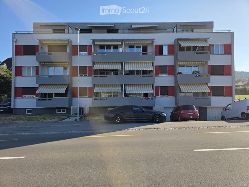 Modern multi-story apartment building with balconies, striped awnings, and several parked cars in front.