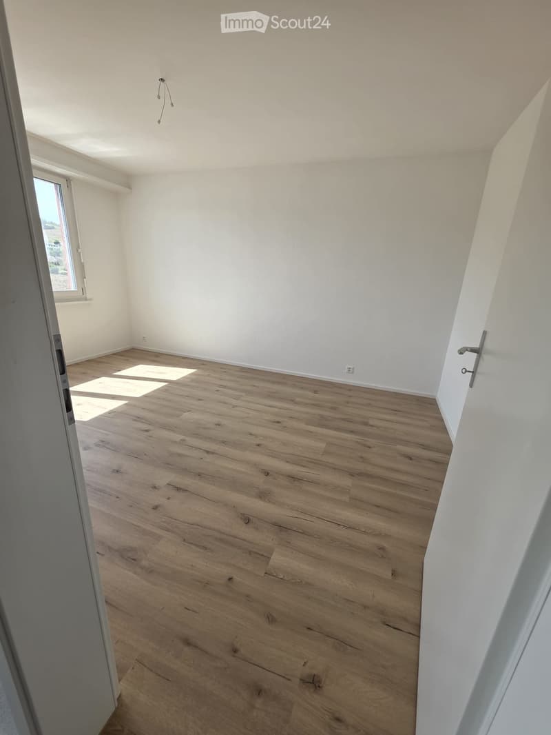 Empty room with light wooden flooring and white walls, featuring a window and a door.
