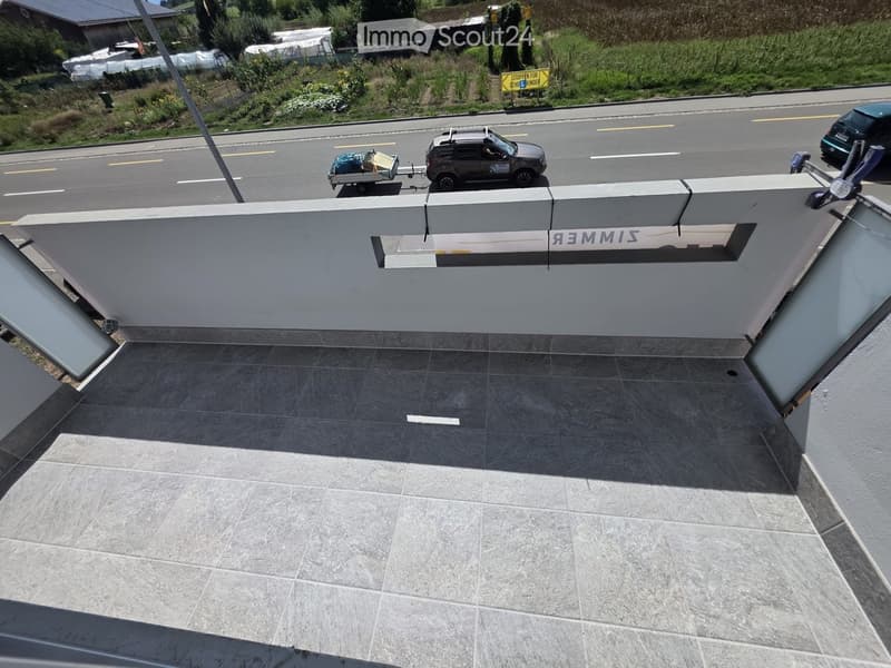 Modern balcony with grey tiled flooring, white railing, and a view of a road with passing vehicles.