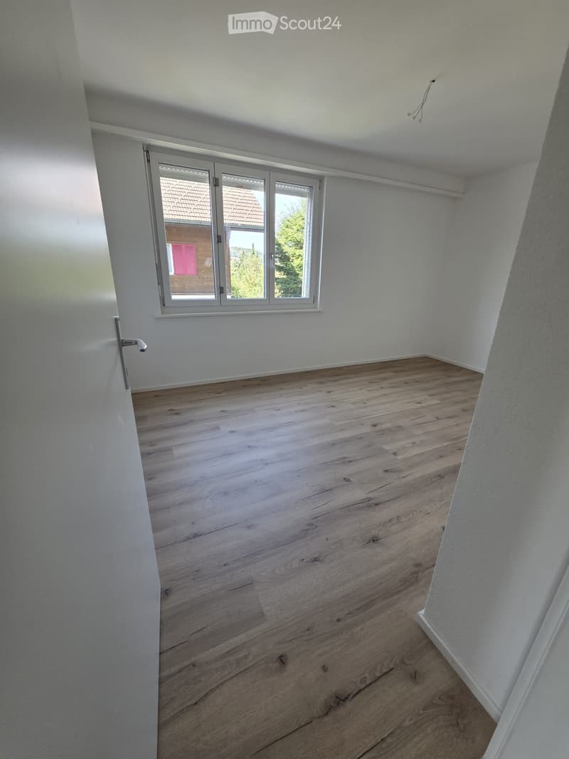 Empty room with wooden flooring, white walls, and a large window with a view of a neighboring building and greenery.