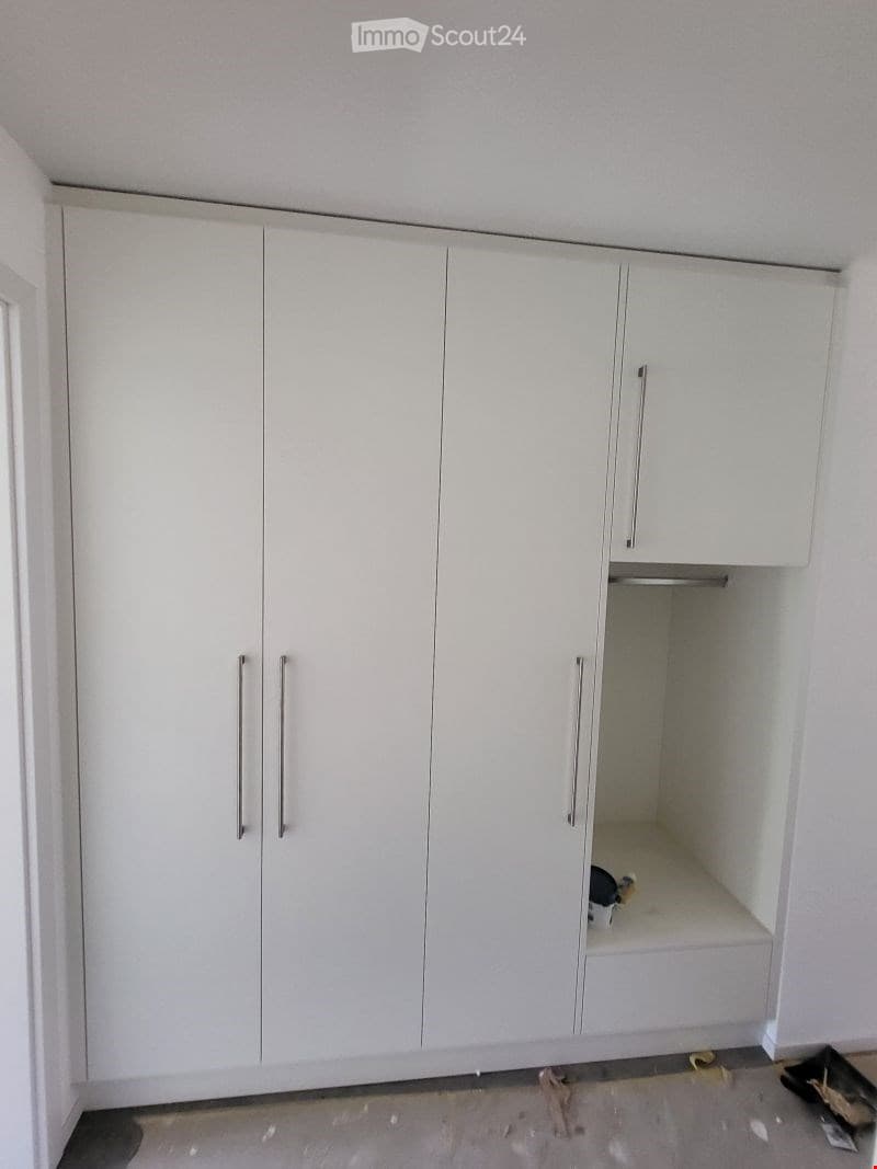 Empty room with white built-in wardrobes and a small open storage space. The floor is unfinished concrete, and there are some construction materials visible.