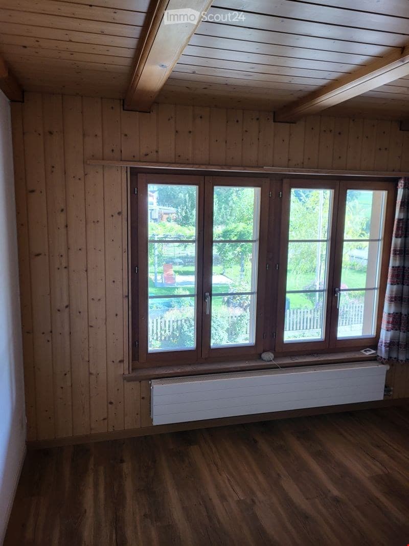Empty room with wooden walls, wooden floor, large window with view of garden, radiator, and exposed wooden beams on the ceiling.