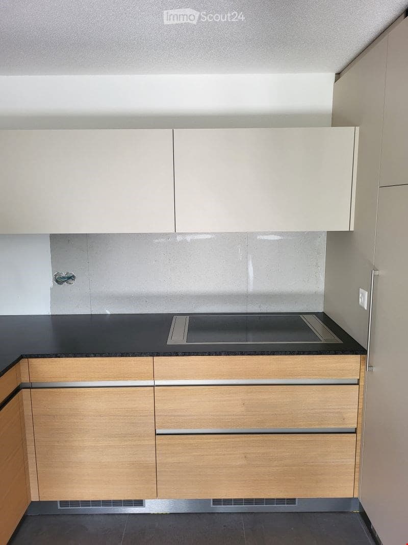 Modern kitchen with light wood cabinets, black countertop, and built-in stove. The walls are grey with a sleek, contemporary design.