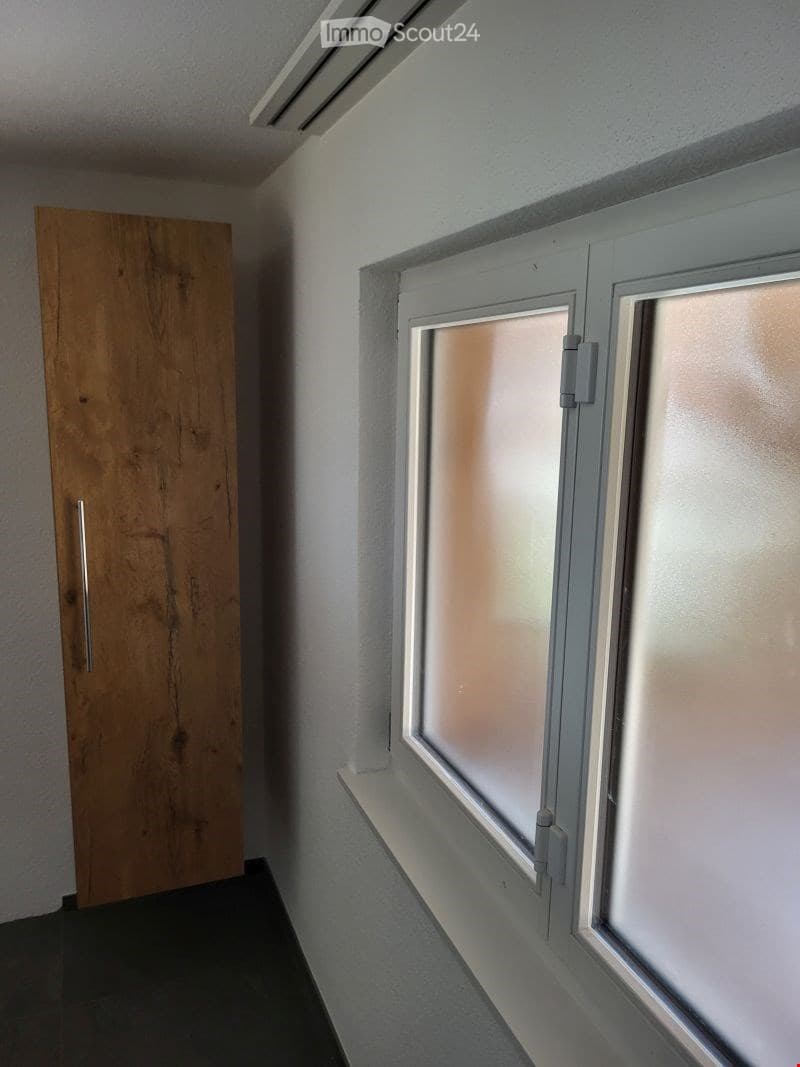 Empty hallway with a wooden door on the left and a window on the right, featuring white walls and a dark floor.