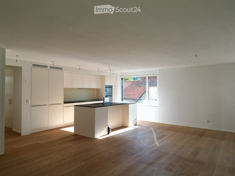 Modern kitchen with light-colored cabinetry, a central island, and wooden flooring. The space is well-lit with large windows offering a view of the roof.