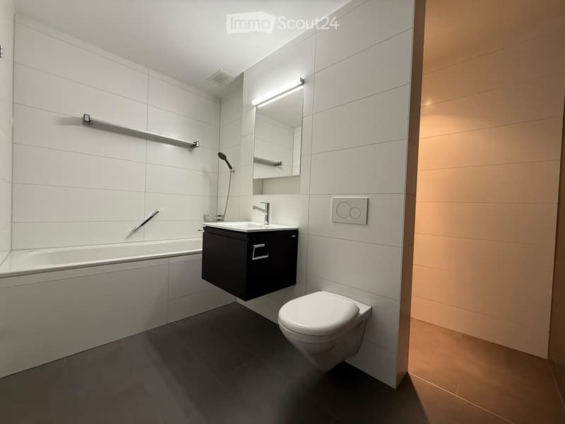 Modern bathroom with white tiles, a bathtub, a sink with a mirror, and a toilet. Features a sleek black vanity cabinet and a built-in showerhead.