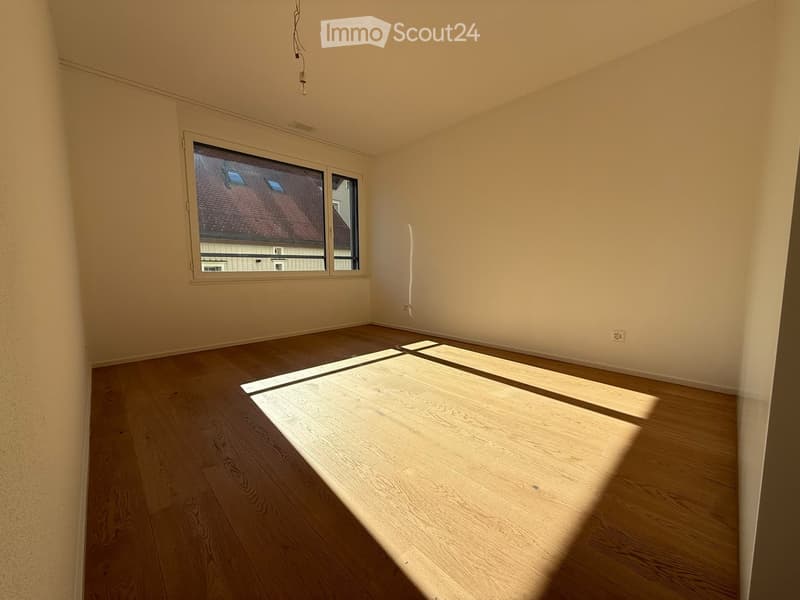 Empty room with wooden flooring and large windows allowing natural light to enter. The walls are painted a light beige color, and there are no visible furnishings or decorations.