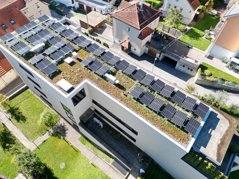 Aerial view of a modern house with a green roof and solar panels, surrounded by neighboring houses and greenery.